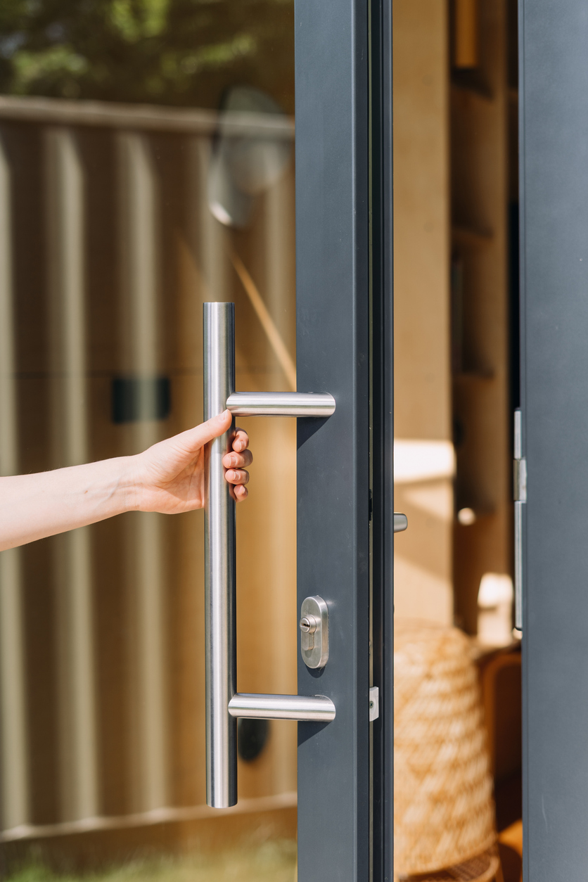 Vertical shot of a woman hand opening a modern front door with a transparent glass panel. The sleek design features a stainless steel handle, enhancing the contemporary look of the home entrance.